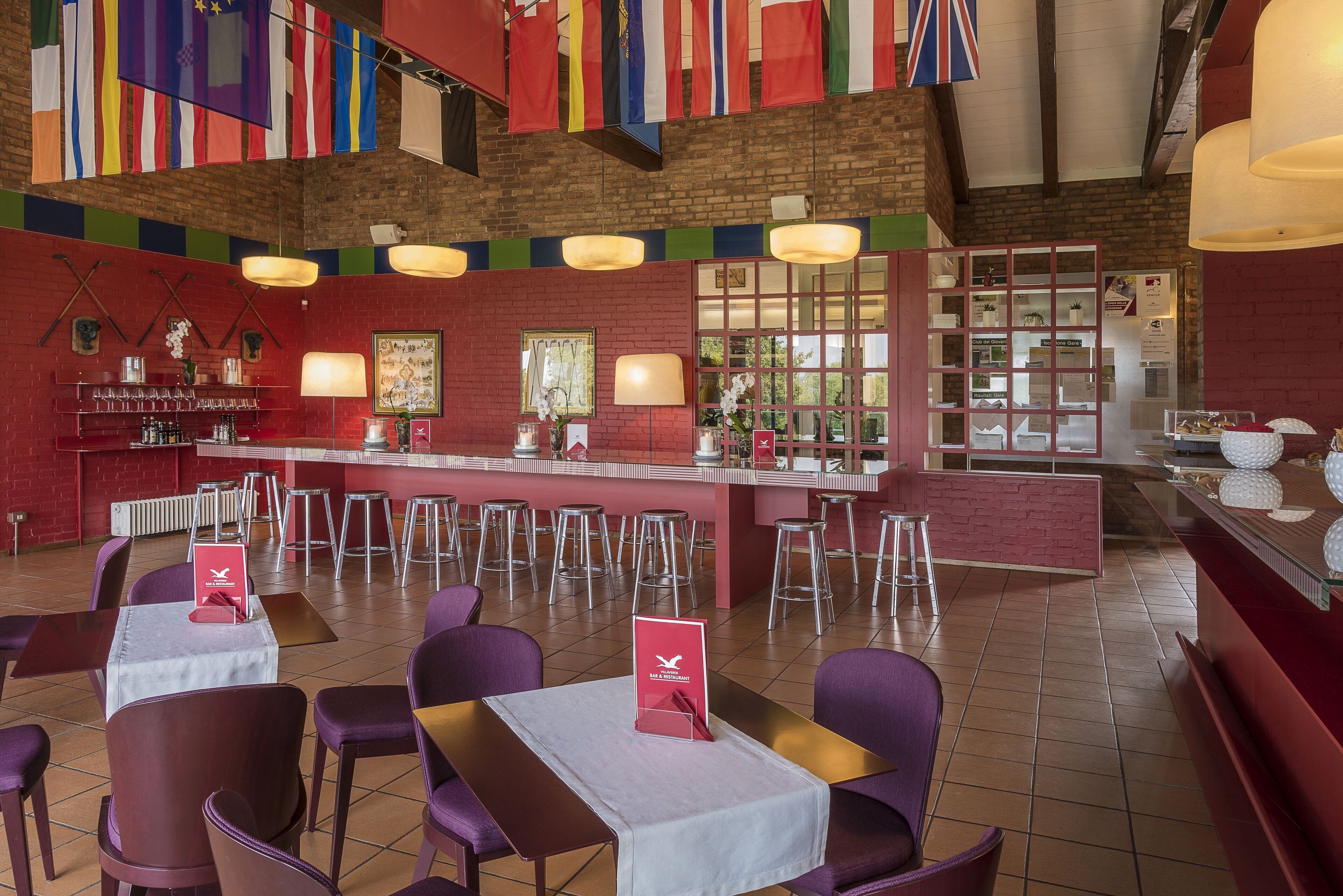 A restaurant with red brick walls, multiple tables and chairs, and a bar area. Flags are hanging from the ceiling.