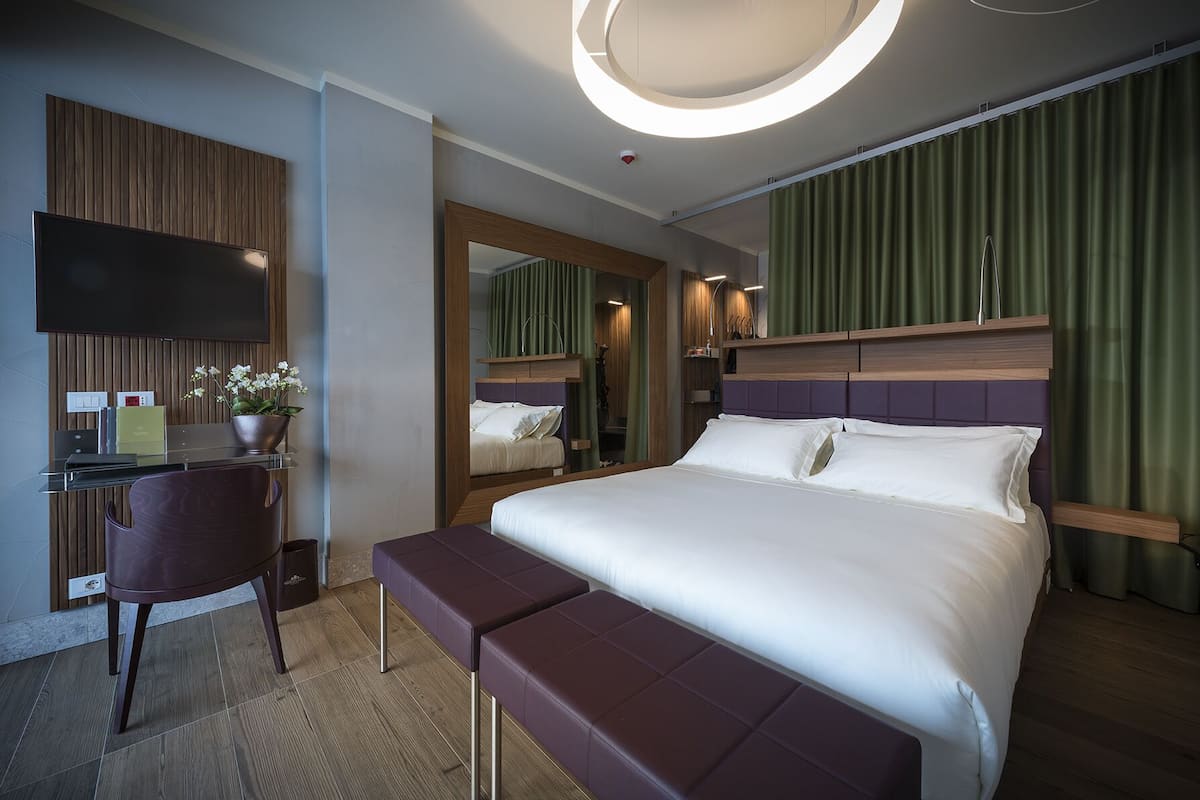 A modern hotel room with a large bed, a desk, a chair, and a mirror.