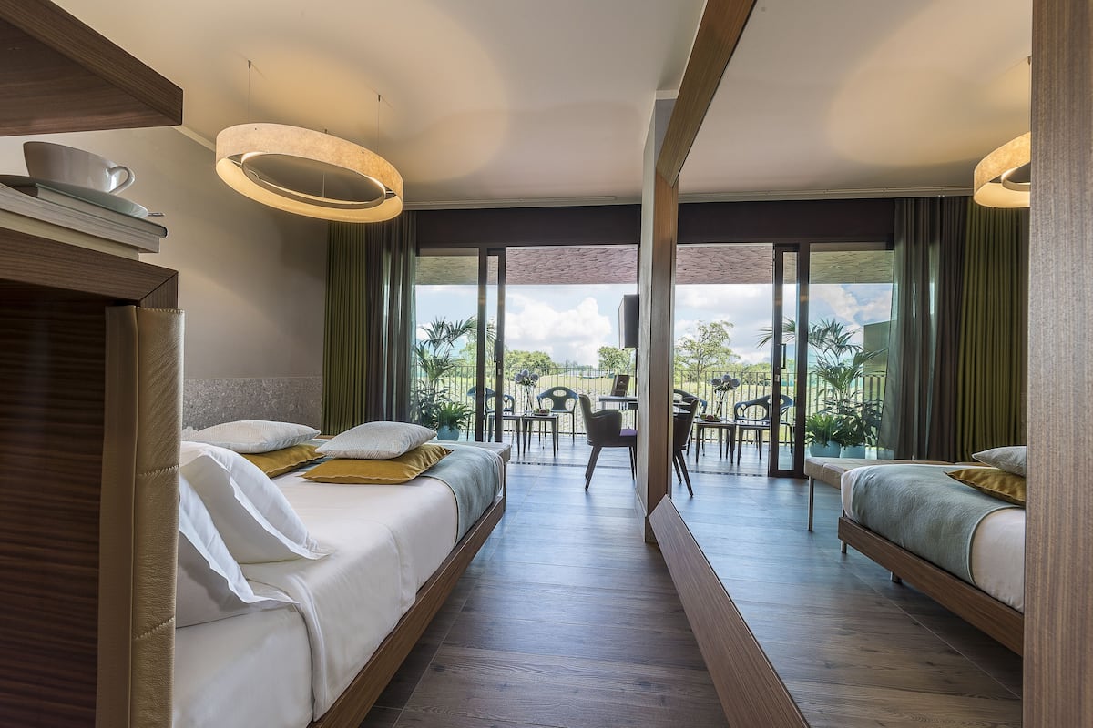 A modern hotel room with two beds, a balcony with a dining area, and large windows offering a view of the outdoors.