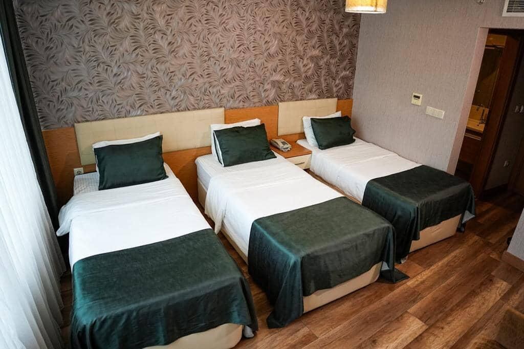Ein Hotelzimmer mit drei Einzelbetten, jedes mit einer grünen Tagesdecke und passenden Kissen. Das Zimmer verfügt über eine gemusterte Tapete und Holzböden.