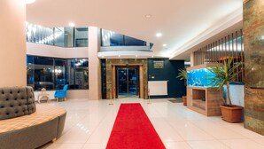 A hotel lobby with a red carpet, a grey sofa, a wooden cabinet, and a large aquarium. - Aksular Hotel (Trabzon)