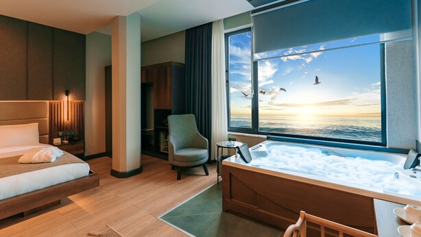 A modern hotel room with a large window, a bed, a chair, a small table, and a bathtub filled with bubbles. - Aksular Hotel (Trabzon)