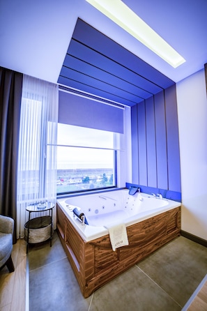 A modern bathroom with a large bathtub, a window with a view, and a small table with a towel. - Aksular Hotel (Trabzon)