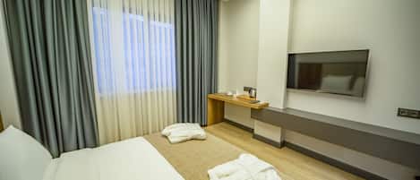 A hotel room with a large bed, a TV mounted on the wall, a small desk with a mirror, and a window with curtains.