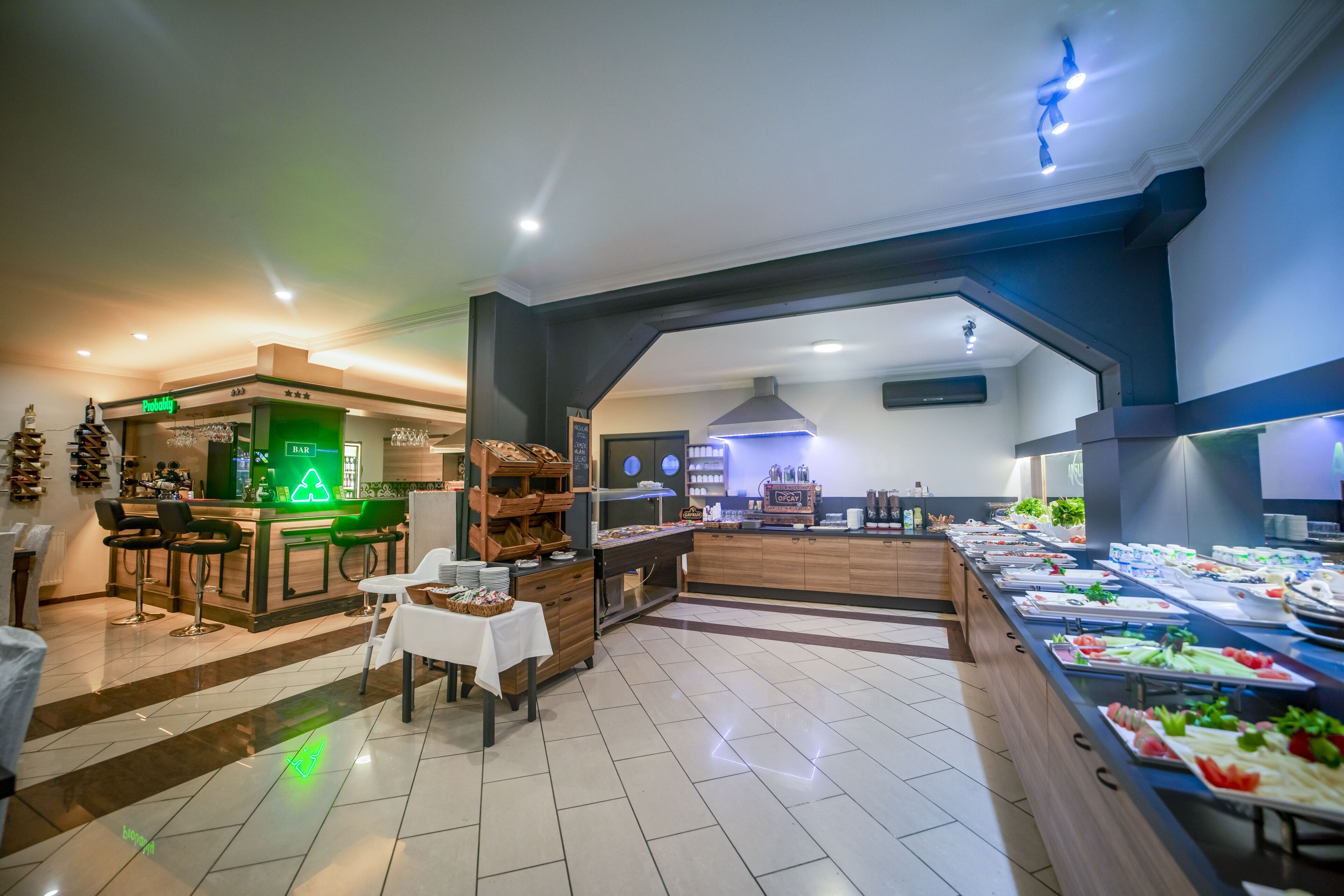 A modern hotel lobby with a bar area, a buffet with various food items, and a seating area with tables and chairs.