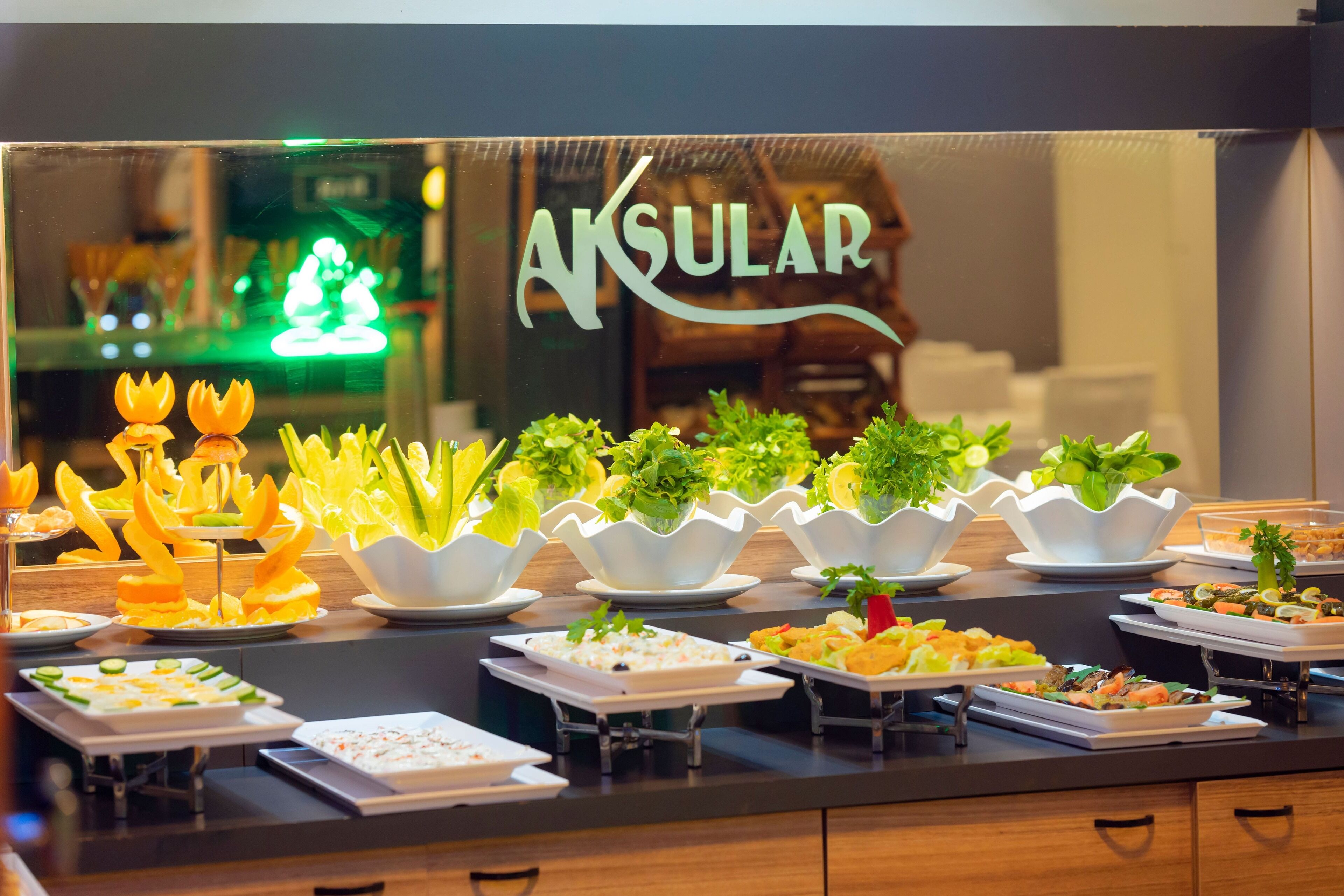 Buffet com diversos pratos e um letreiro ao fundo com a inscrição 'AKSULAR'.