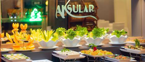Buffet com diversos pratos e um letreiro ao fundo com a inscrição 'AKSULAR'.