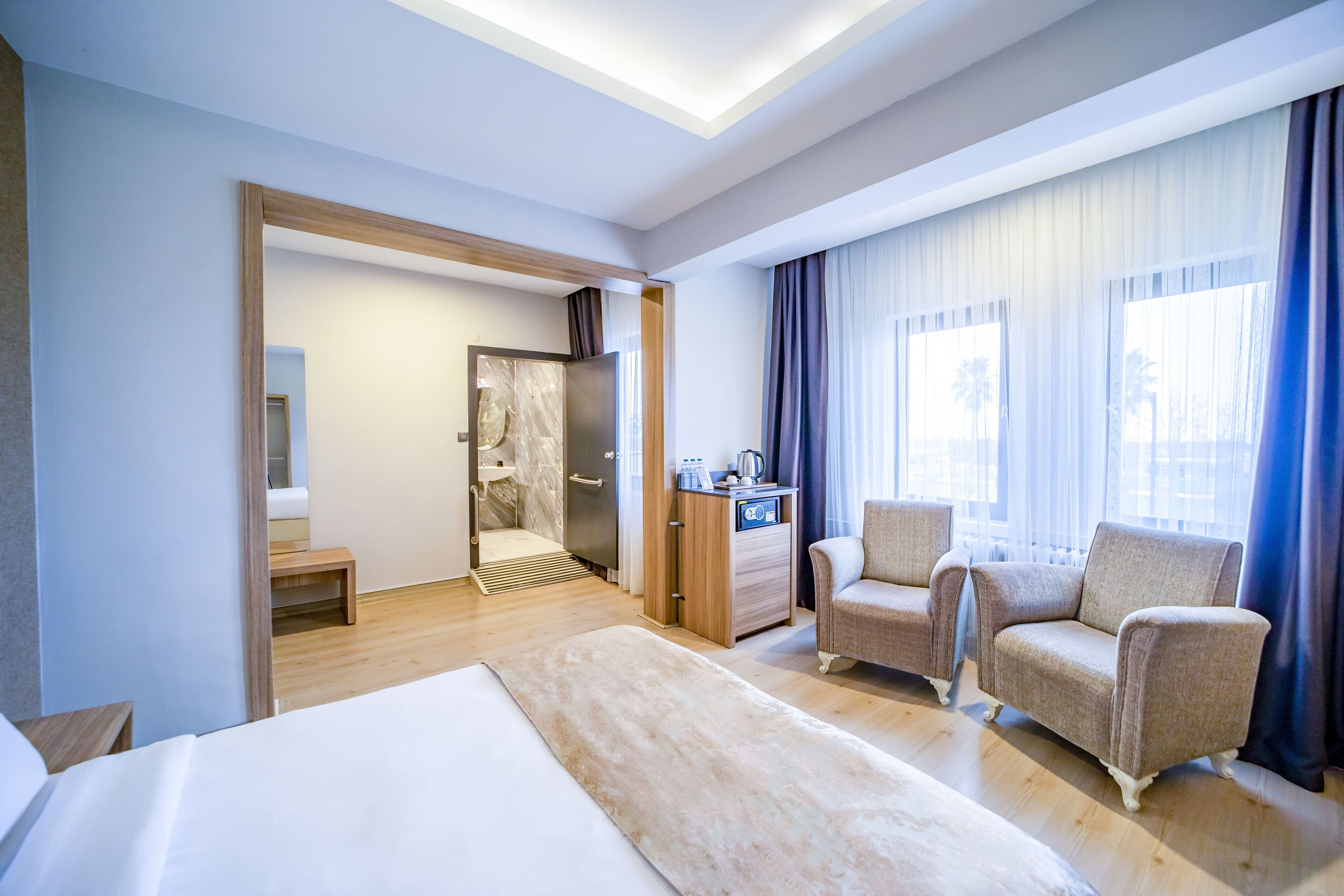 A hotel room with a bed, two armchairs, a small table, a shower area, and a view of palm trees through the window.