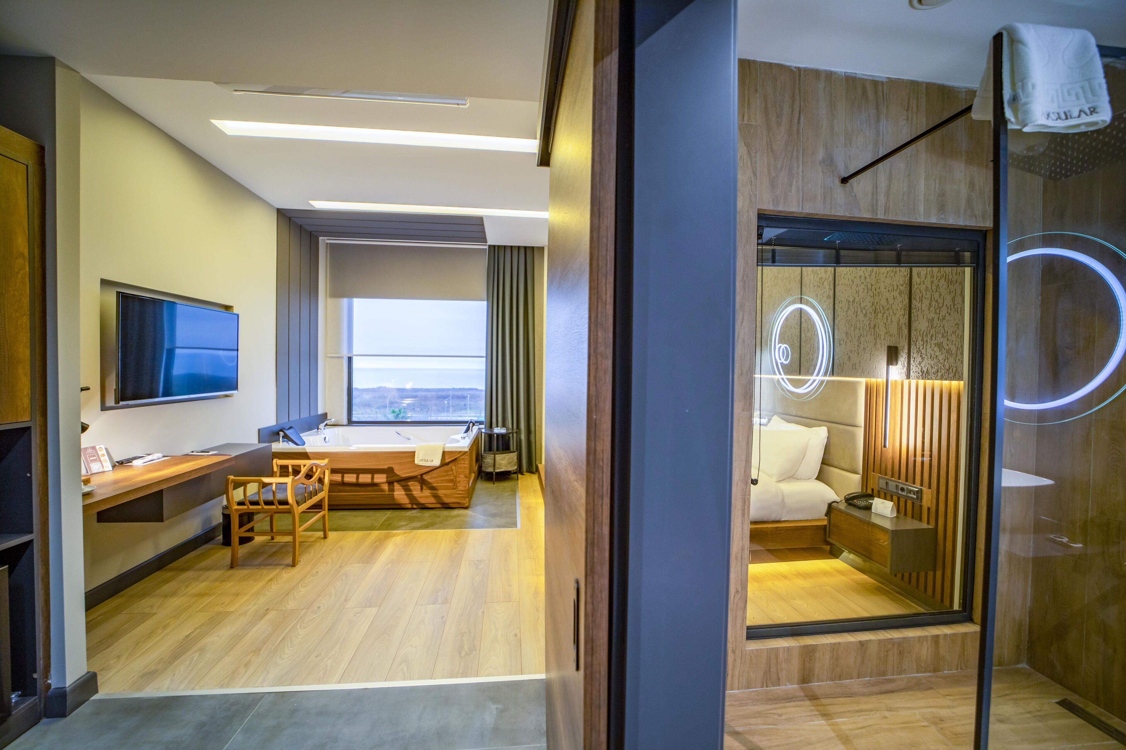A modern hotel room with a wooden floor, a desk with a flat-screen TV, a bed with a headboard, and a view of the city.