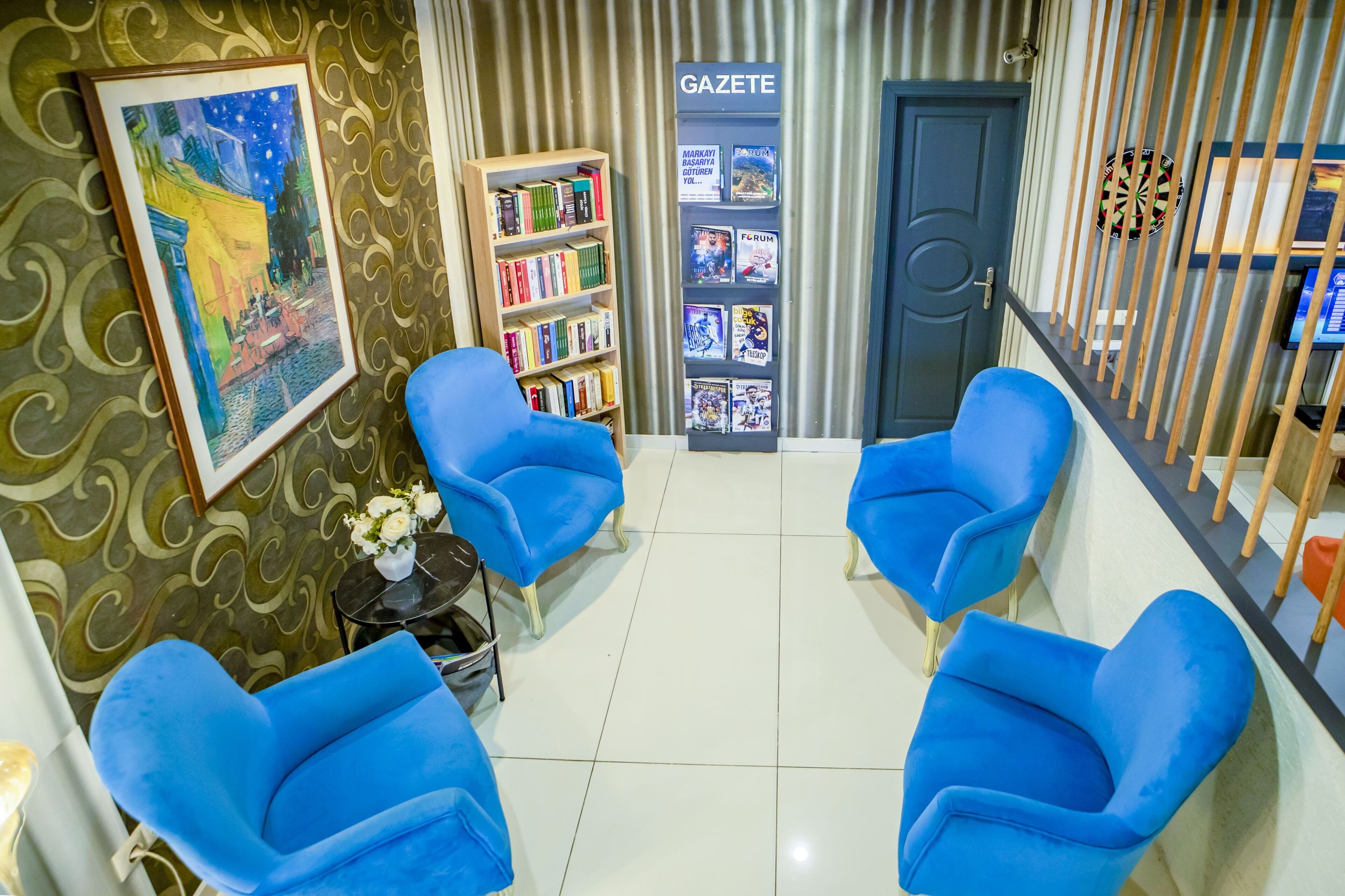 A lobby with blue armchairs, a bookshelf, a framed painting, and a dartboard.