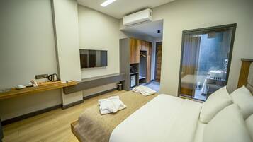 A modern hotel room with a large bed, a flat-screen TV mounted on the wall, a wooden side table with a kettle, and a view of the hotel's interior through a glass door.