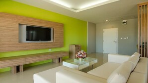 A modern living room with a large flat-screen TV mounted on a wooden panel, a glass coffee table with a vase of flowers, and a beige sofa. - The Lake Hotel (Khon Kaen)