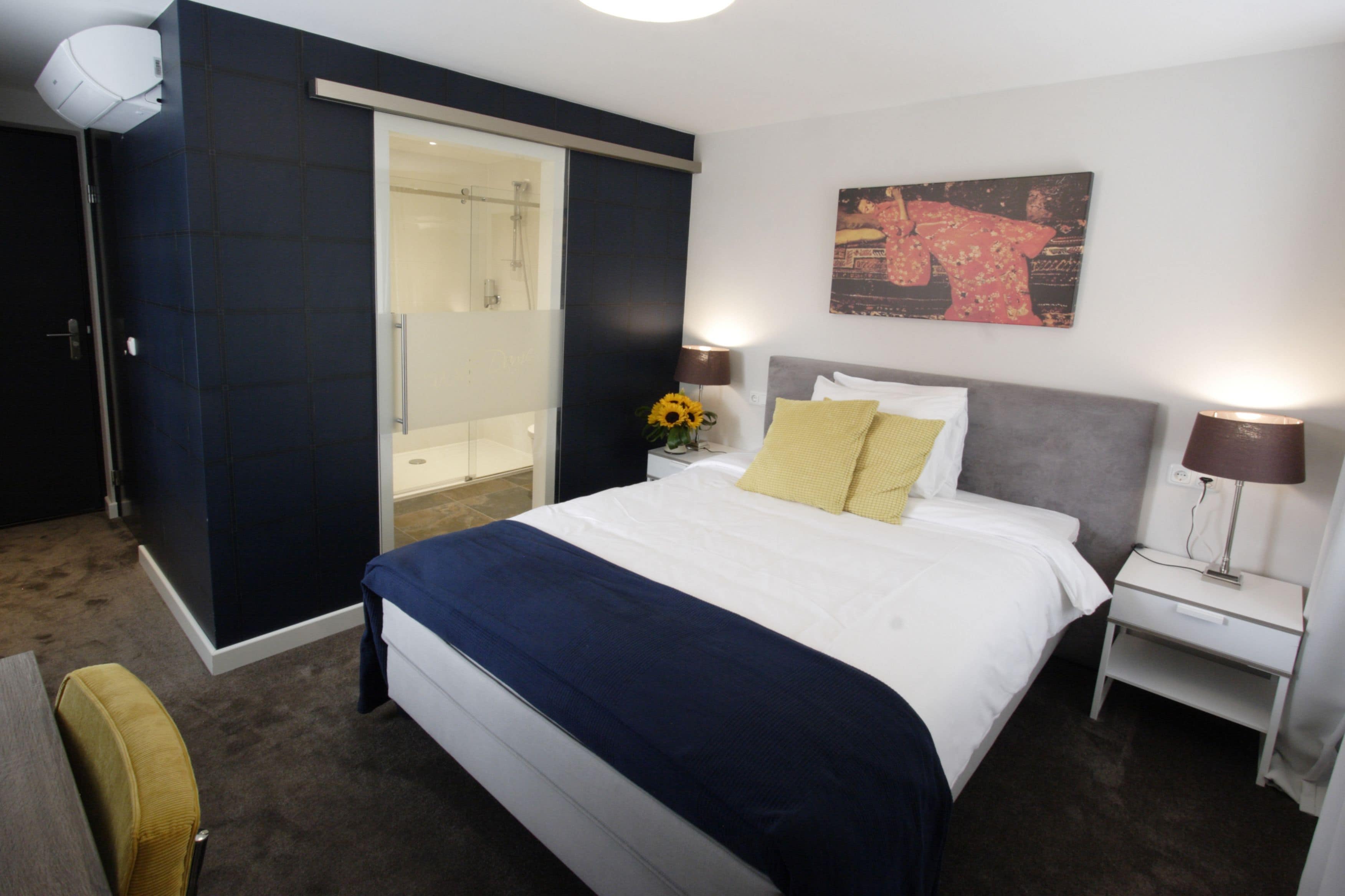 A hotel room with a large bed, a nightstand with lamps, a wall-mounted painting, and a sliding door leading to a bathroom.