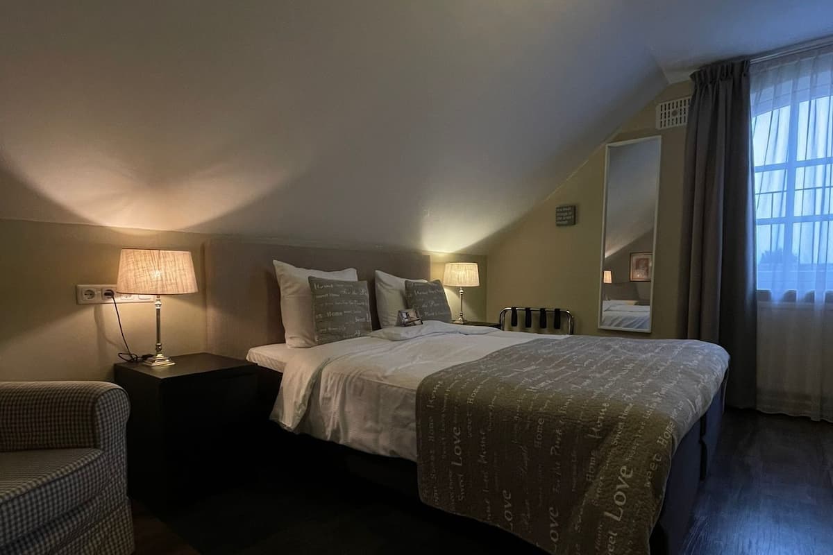 A bedroom with a large bed, a bedside table, a lamp, a chair, and a window with curtains.