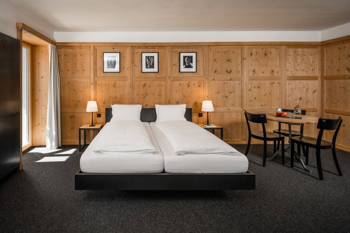 A double bed with two nightstands, a desk with a chair, and wooden panel walls with framed pictures.