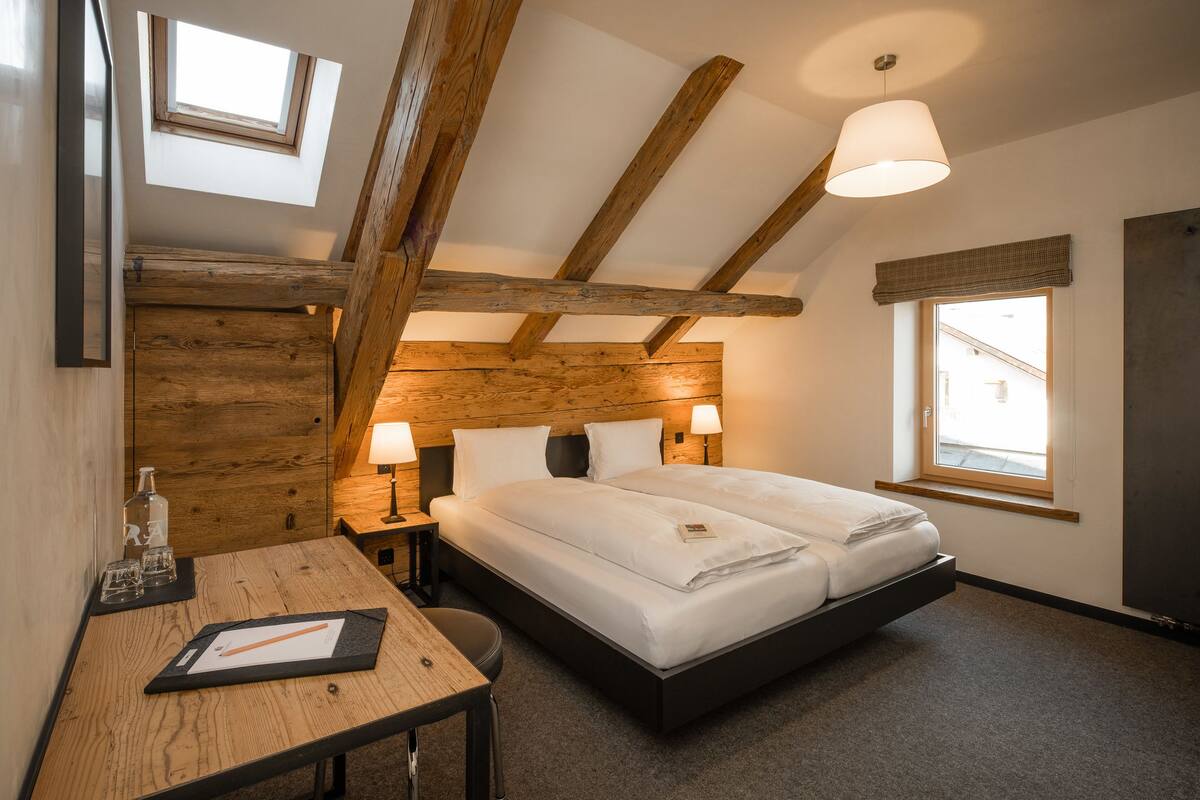 A bedroom with a loft-style bed, wooden beams, a desk with a chair, and two bedside lamps.