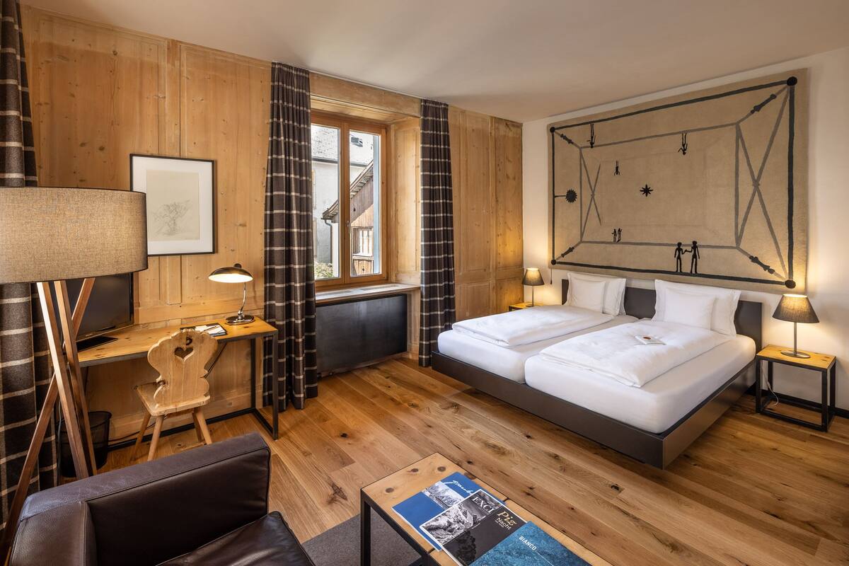 A hotel room with a wooden floor, a bed, a desk, a chair, a lamp, and a large framed artwork on the wall.