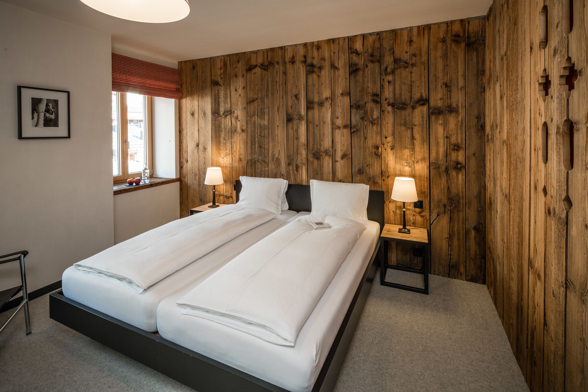 Una cama doble con sábanas blancas, dos mesitas de noche con lámparas, un panel de madera en la pared, un cuadro enmarcado y una ventana con un adorno rojo.