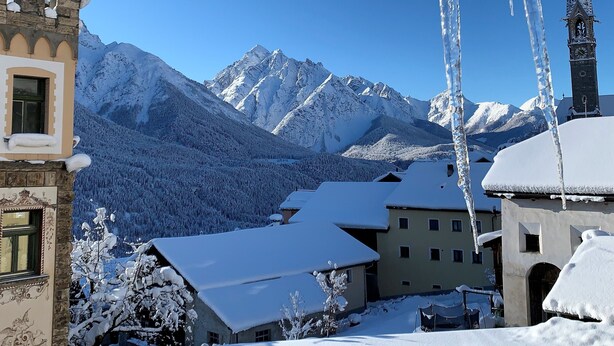 Un pueblo cubierto de nieve en la montaña, con arquitectura tradicional, incluyendo una torre de iglesia y un edificio con tejado nevado.