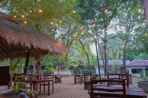 Un área de estar al aire libre con estructuras de techo de paja y mobiliario de madera, rodeada de árboles y luces colgantes.