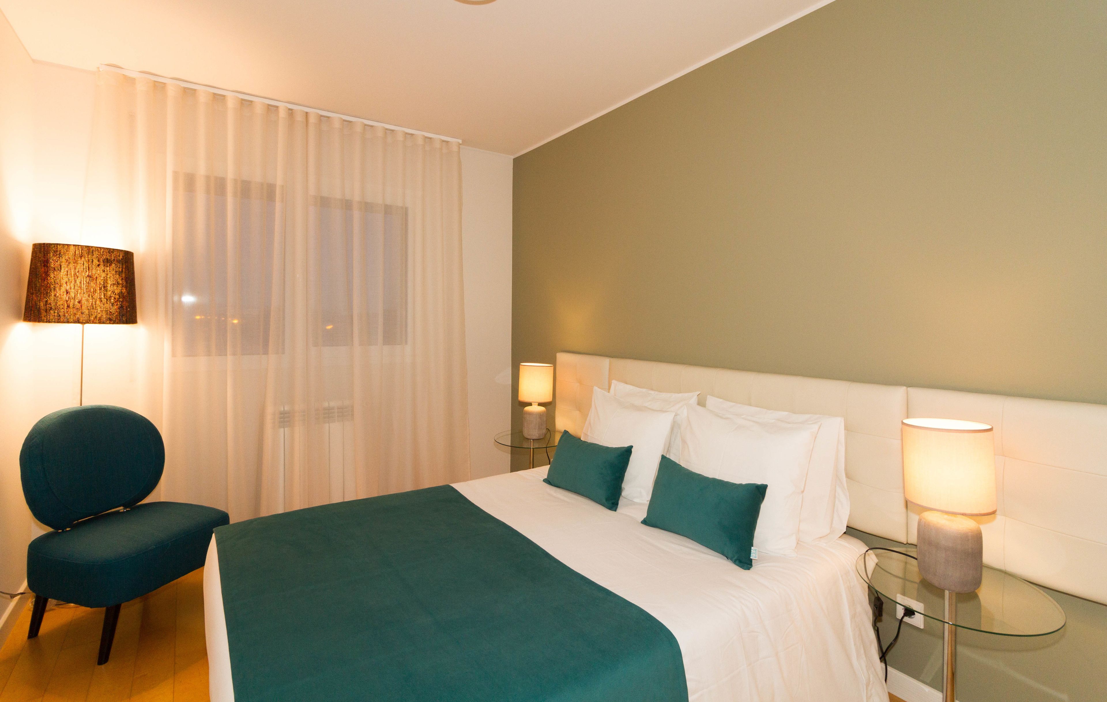 A hotel room with a bed, a chair, a bedside table with a lamp, and a window with sheer curtains.