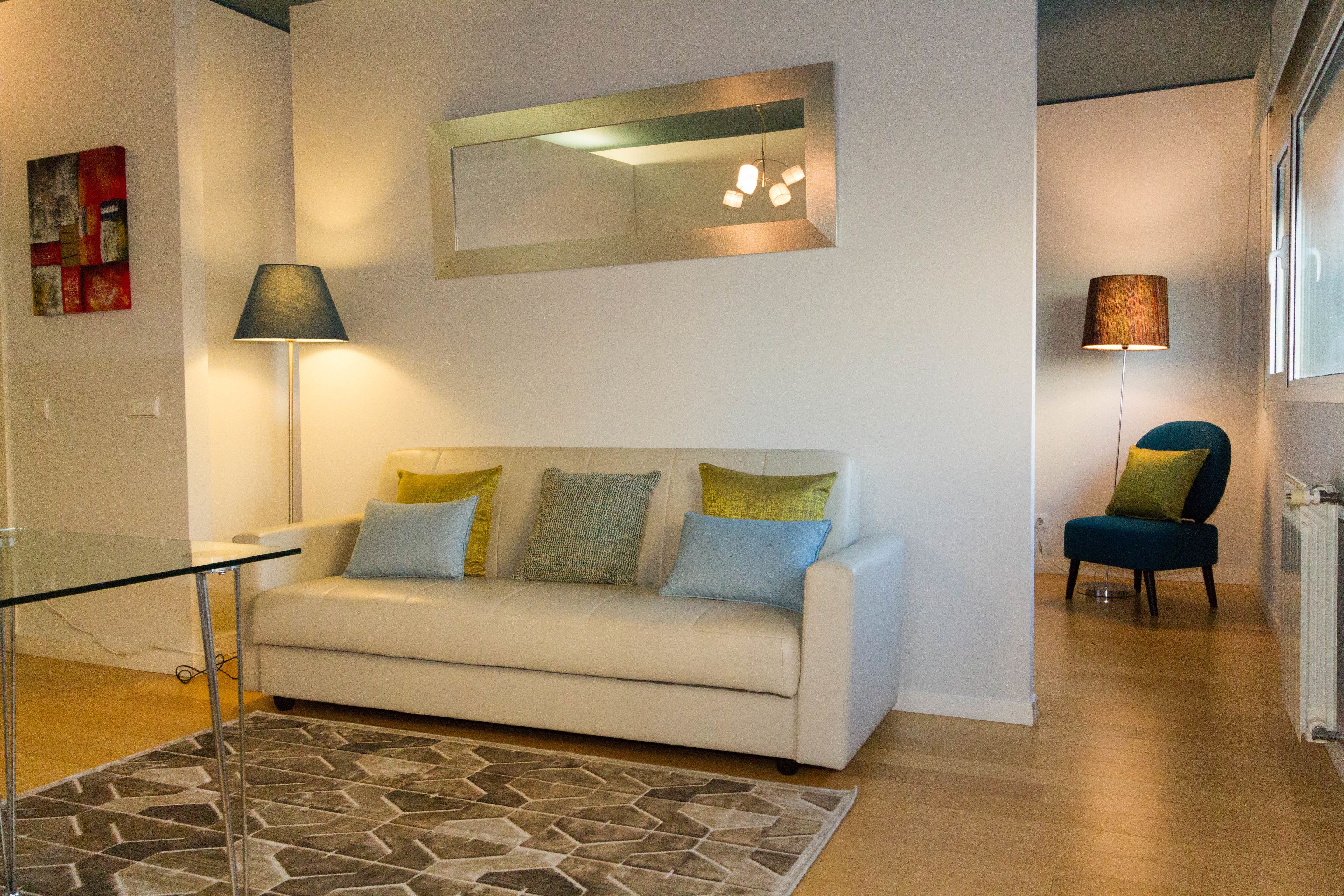 Ein Wohnzimmer mit einem weißen Sofa, einem Glastisch, einem Teppich mit Muster, einem Spiegel und zwei Lampen.