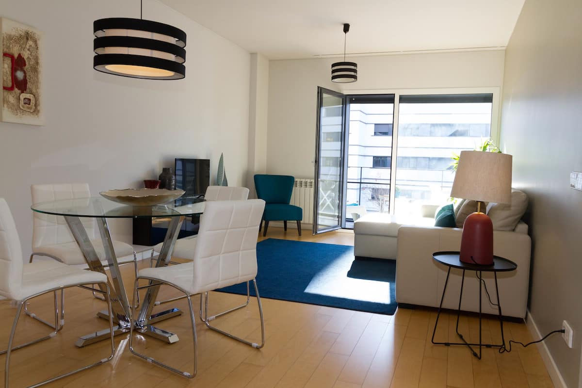 A modern living room with a glass dining table, white sofas, and a blue rug.
