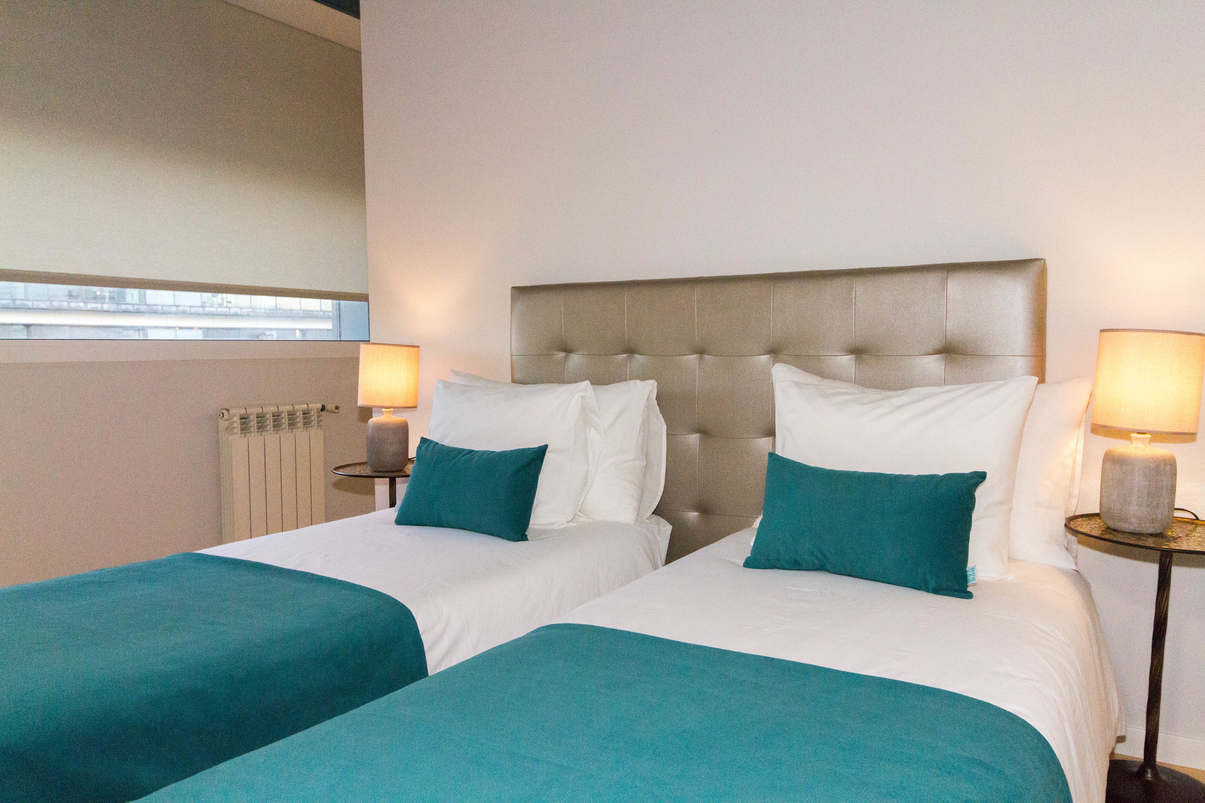 Une chambre d’hôtel avec deux lits, chacun agrémenté de draps blancs et de coussins décoratifs de couleur turquoise. On y trouve deux tables de chevet avec des lampes et des bougies.