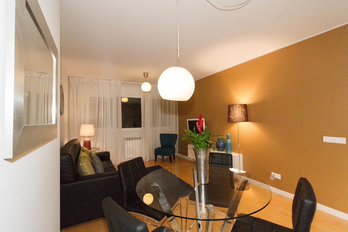 A modern living room with a glass dining table, black sofas, a blue armchair, and a vase with flowers.