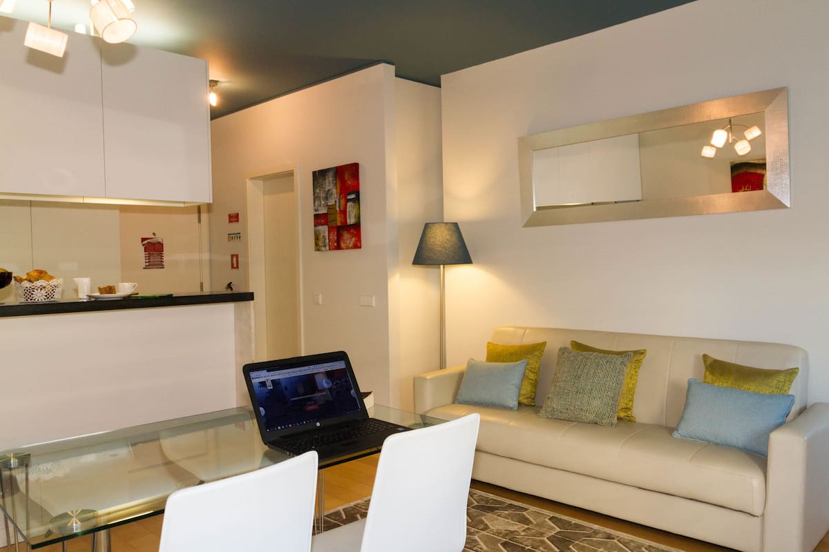 A modern living room with a glass dining table, a laptop, a white sofa with colorful cushions, and a kitchen area with white cabinets and a red fire extinguisher.
