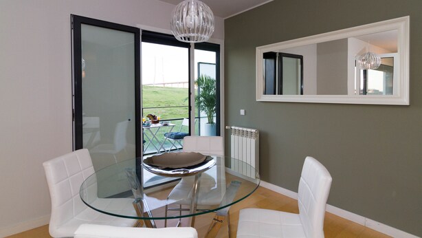 A modern dining area with a glass table and white chairs, a large window with a view of the outdoors, and a hanging light fixture. - Oriente Rio Apartments by APT IIN - Parque das Nações (Lisbon)