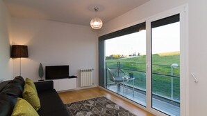 A modern living room with a black sofa, a television, a patterned rug, and a large sliding glass door leading to a balcony with a view of a green field. - Oriente Rio Apartments by APT IIN - Parque das Nações (Lisbon)