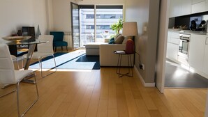 A modern living room with a glass dining table, a sofa, and a coffee table. There is a kitchen area with cabinets and a stove. - Oriente Rio Apartments by APT IIN - Parque das Nações (Lisbon)