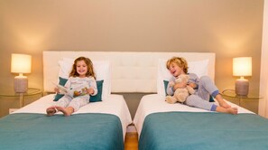 Zwei Kinder in Pyjamas liegen auf einzelnen Betten in einem Hotelzimmer, jedes mit einer Lampe auf einem Nachttisch.