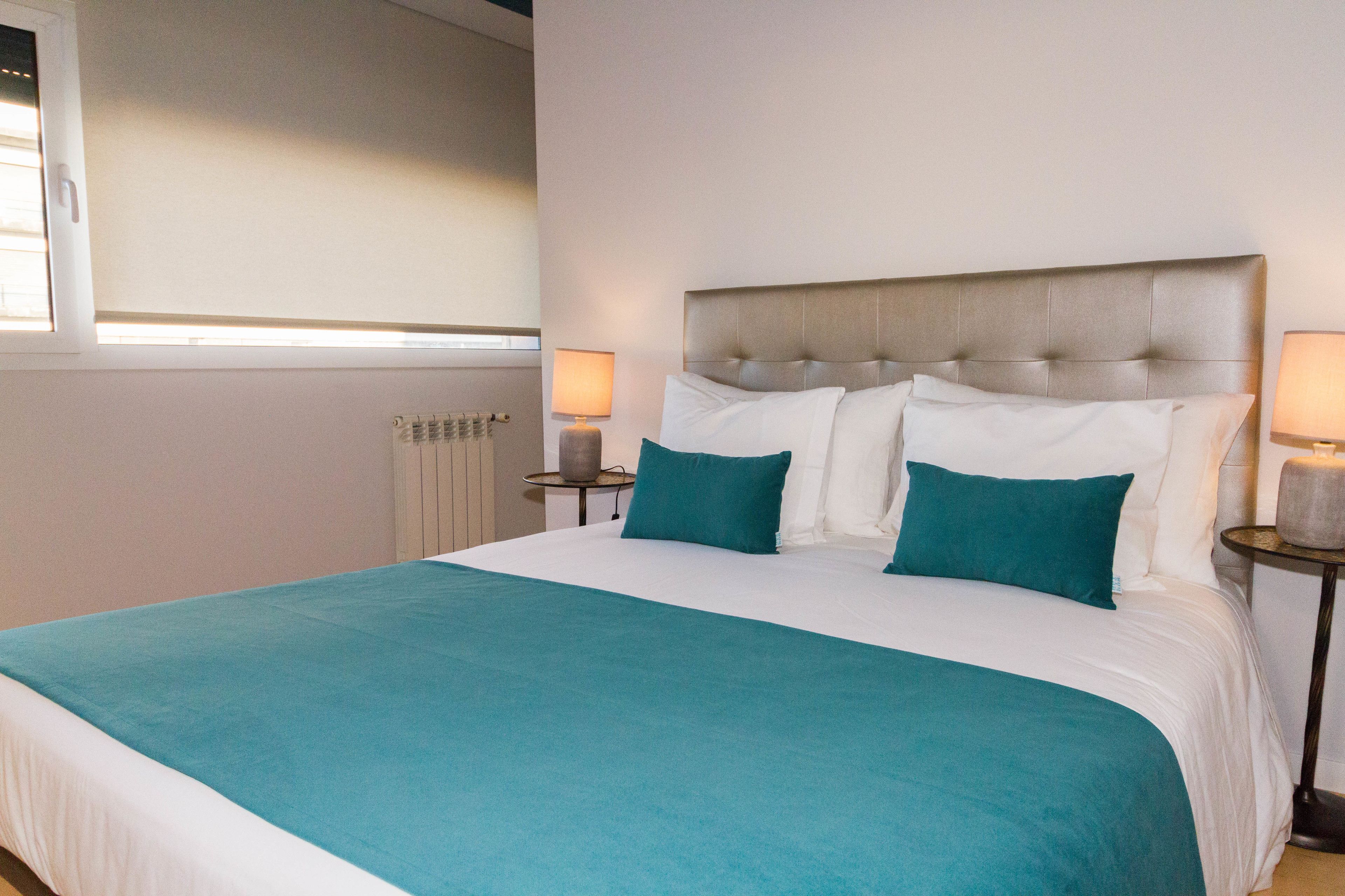A neatly made bed with a teal bedspread and white pillows, two bedside tables with lamps, and a radiator on the wall.