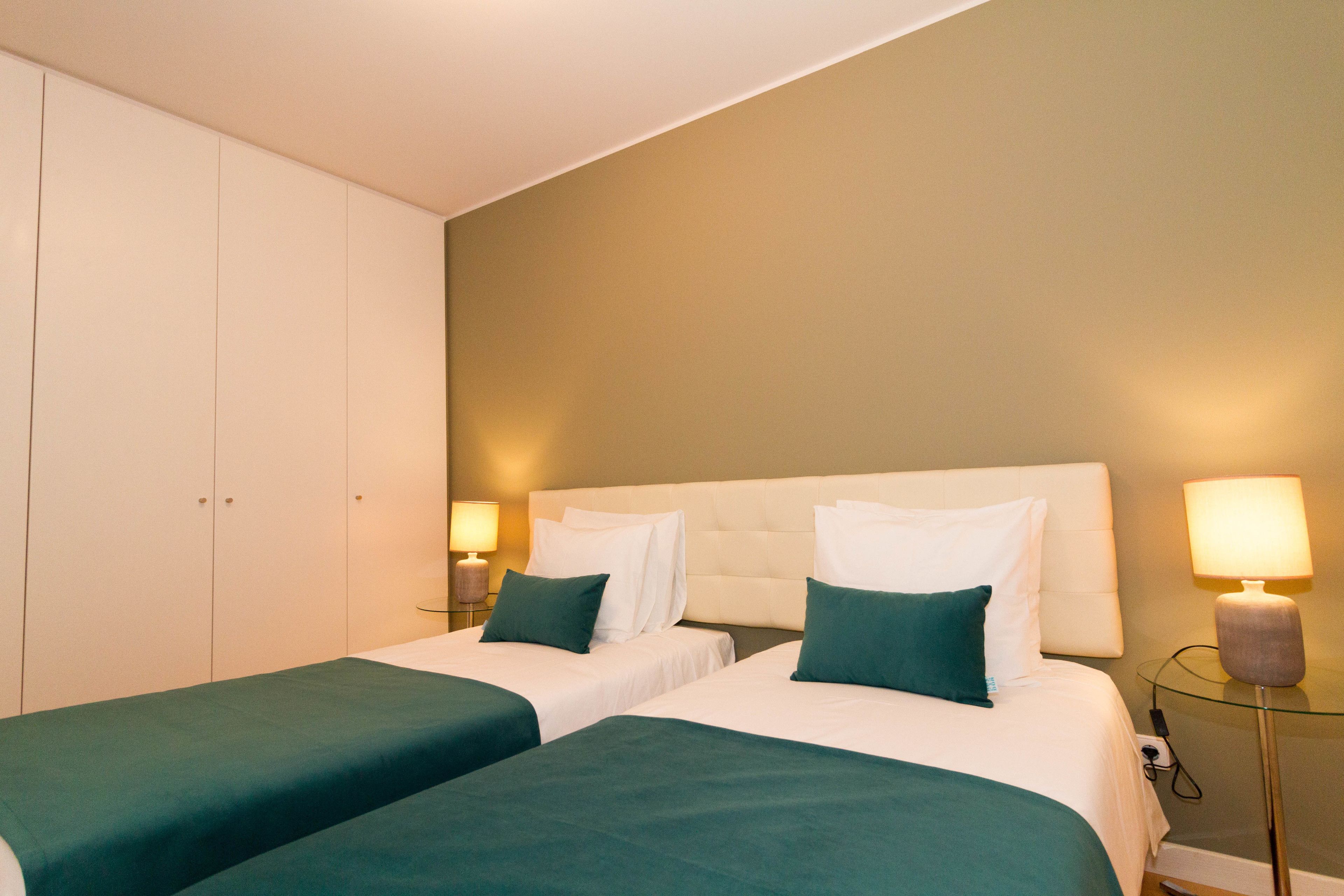 A hotel room with two beds, a white headboard, green bedding, a bedside lamp, and a wardrobe.