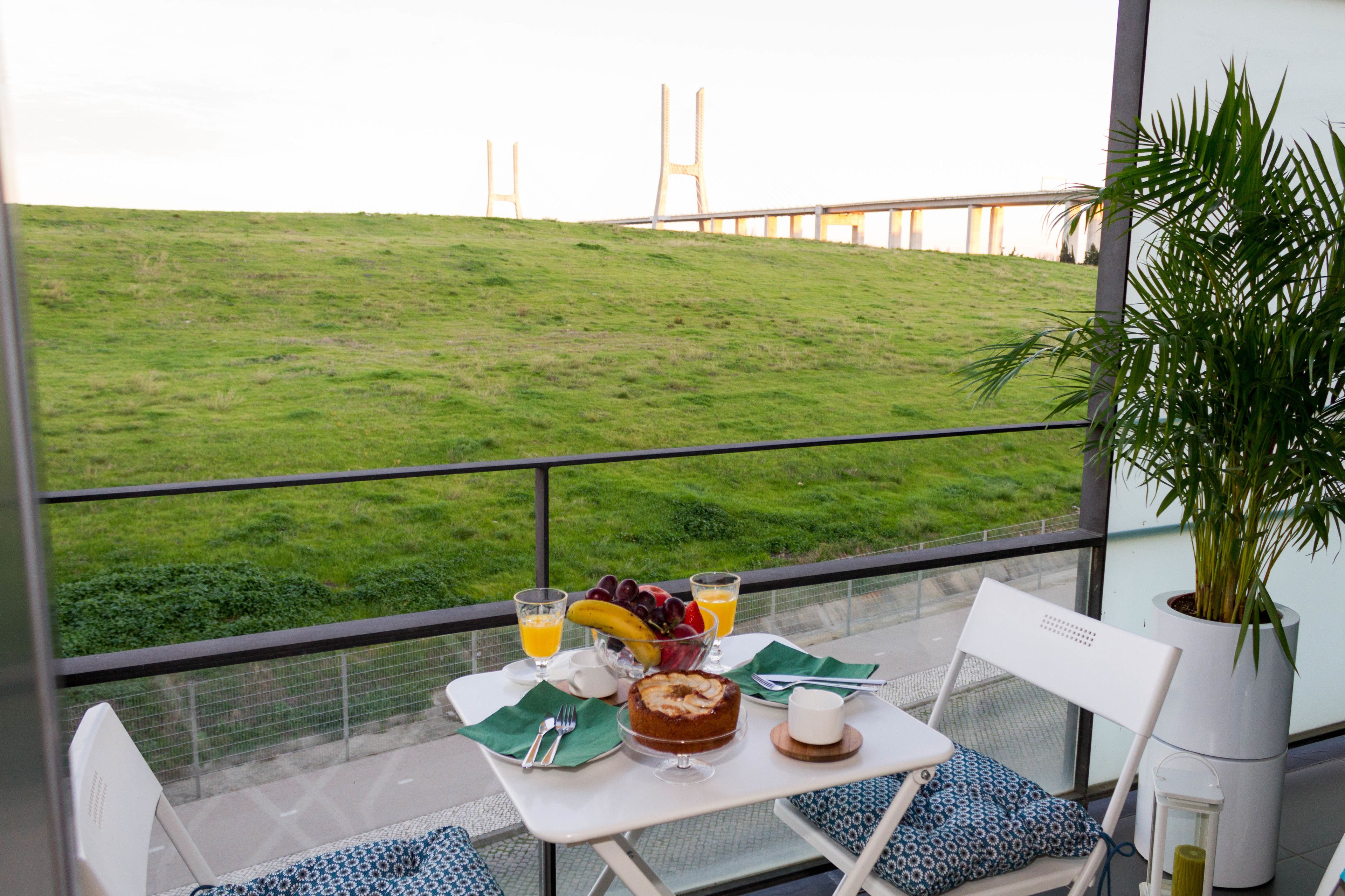 A balcony with a table set for breakfast, a view of a grassy hill, and a bridge in the distance.