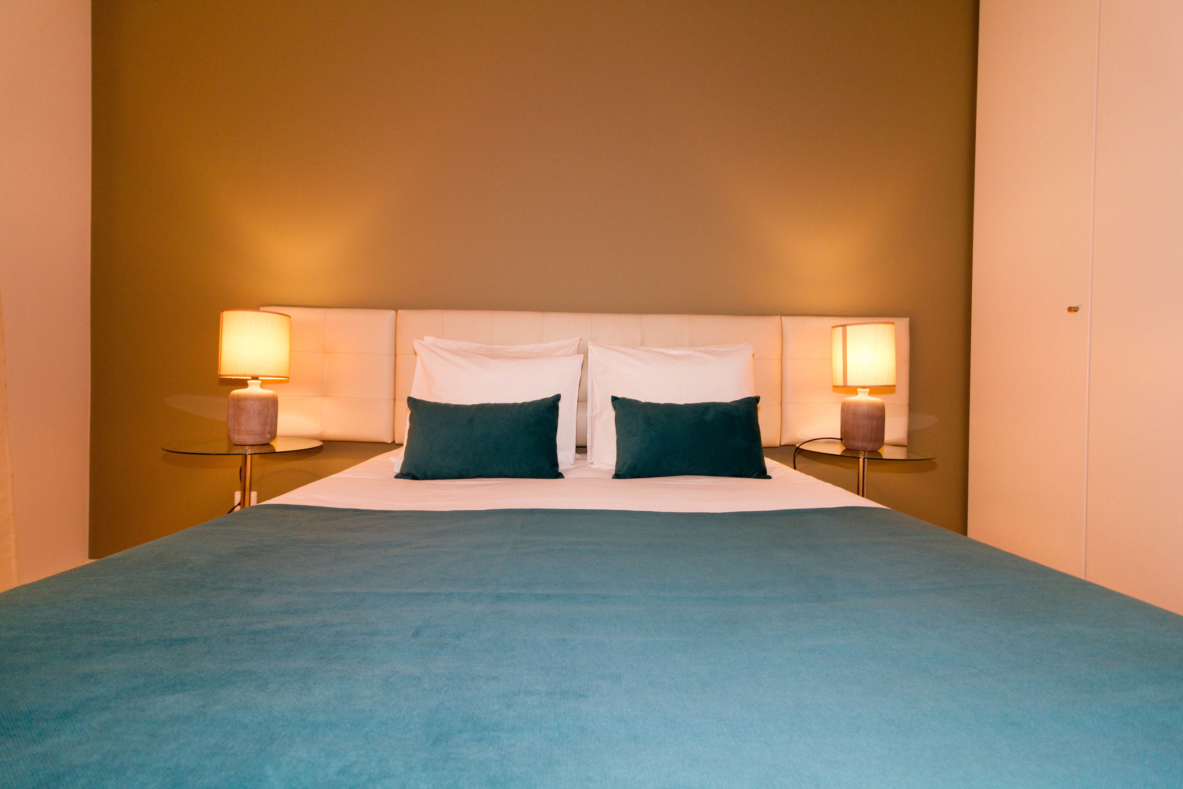 Een opgemaakt bed met een blauw sprei, twee donkere kussens en twee bedlampjes.