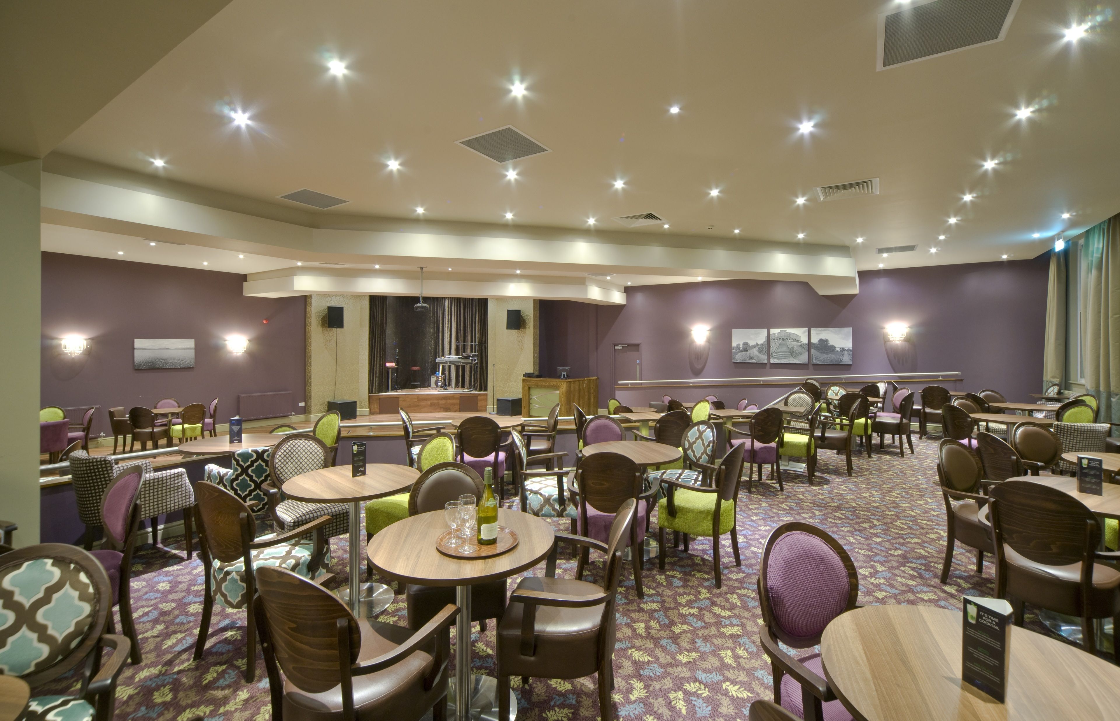 A hotel dining area with round tables, chairs, and a stage for presentations.