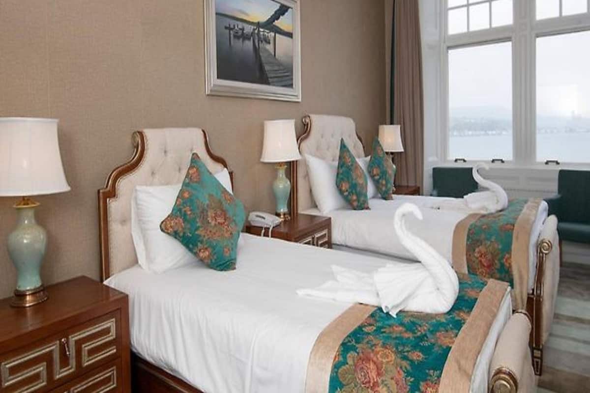A hotel room with two beds, each with a white bedspread and matching headboards, flanked by nightstands with lamps. A framed picture hangs above the beds, and a window with curtains provides a view of the outside.