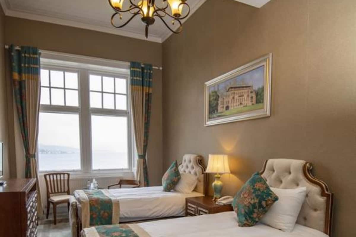 A hotel room with two beds, a chandelier, a desk, and a window with curtains.