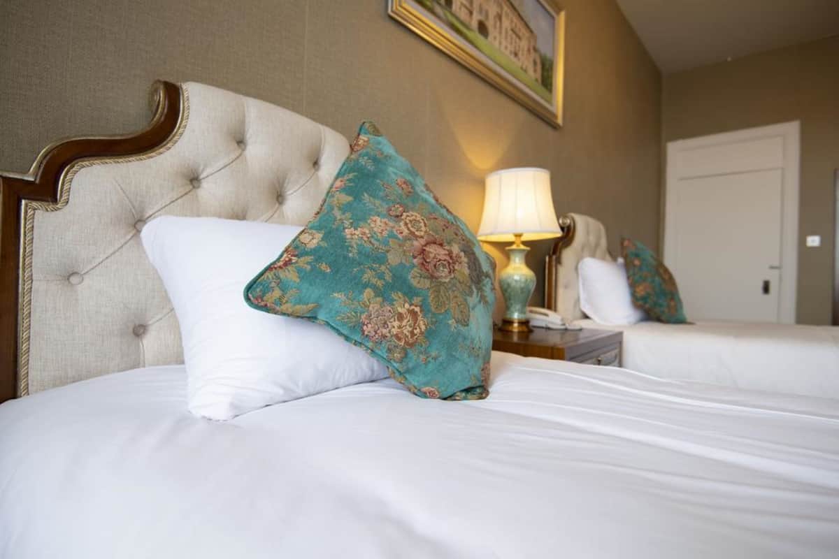 A hotel room with a tufted headboard, a bed with white bedding, a teal floral pillow, and a bedside lamp.