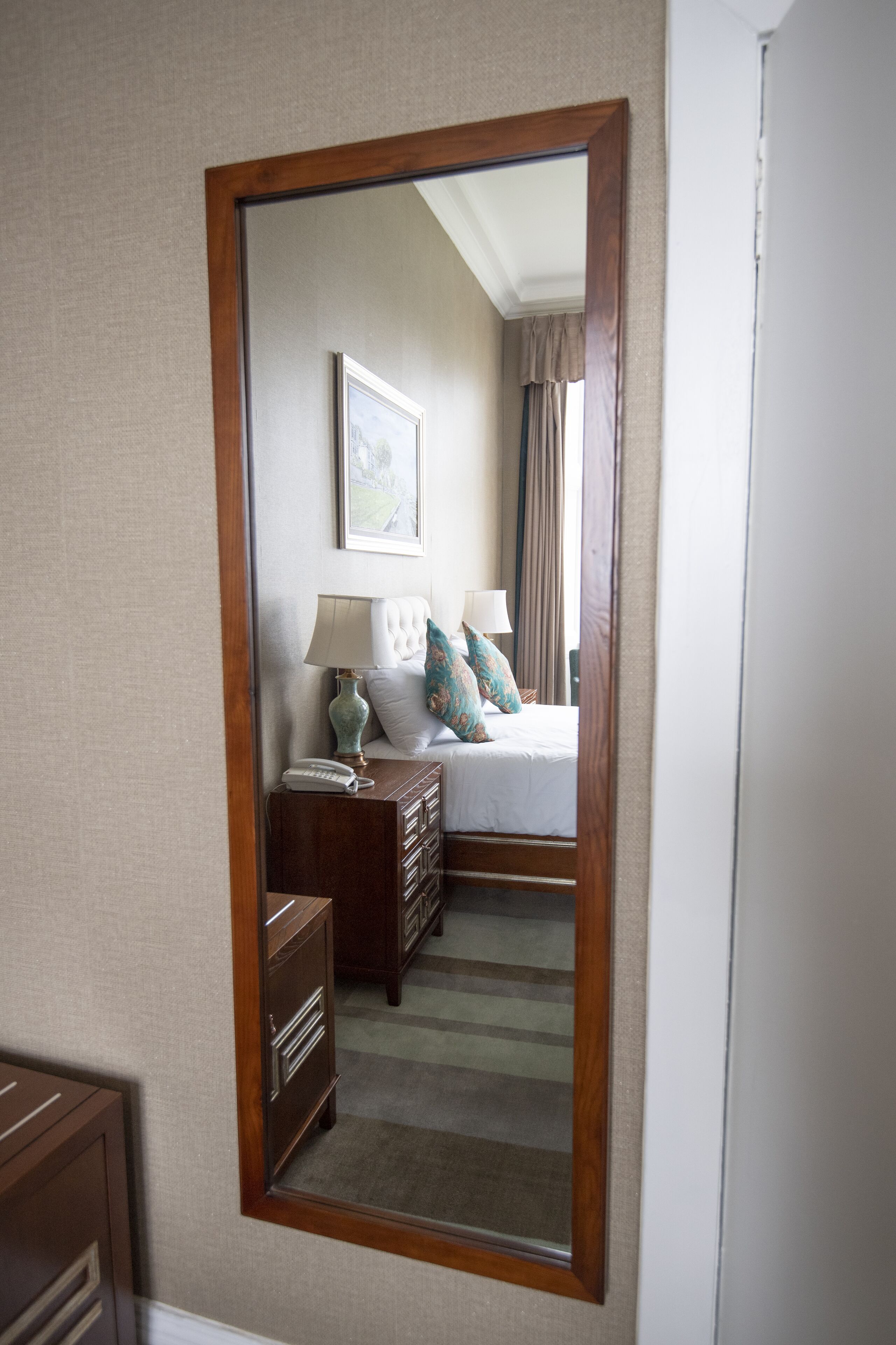 A hotel room with a large mirror, a bed with white linens, a nightstand with a lamp, and a dresser.