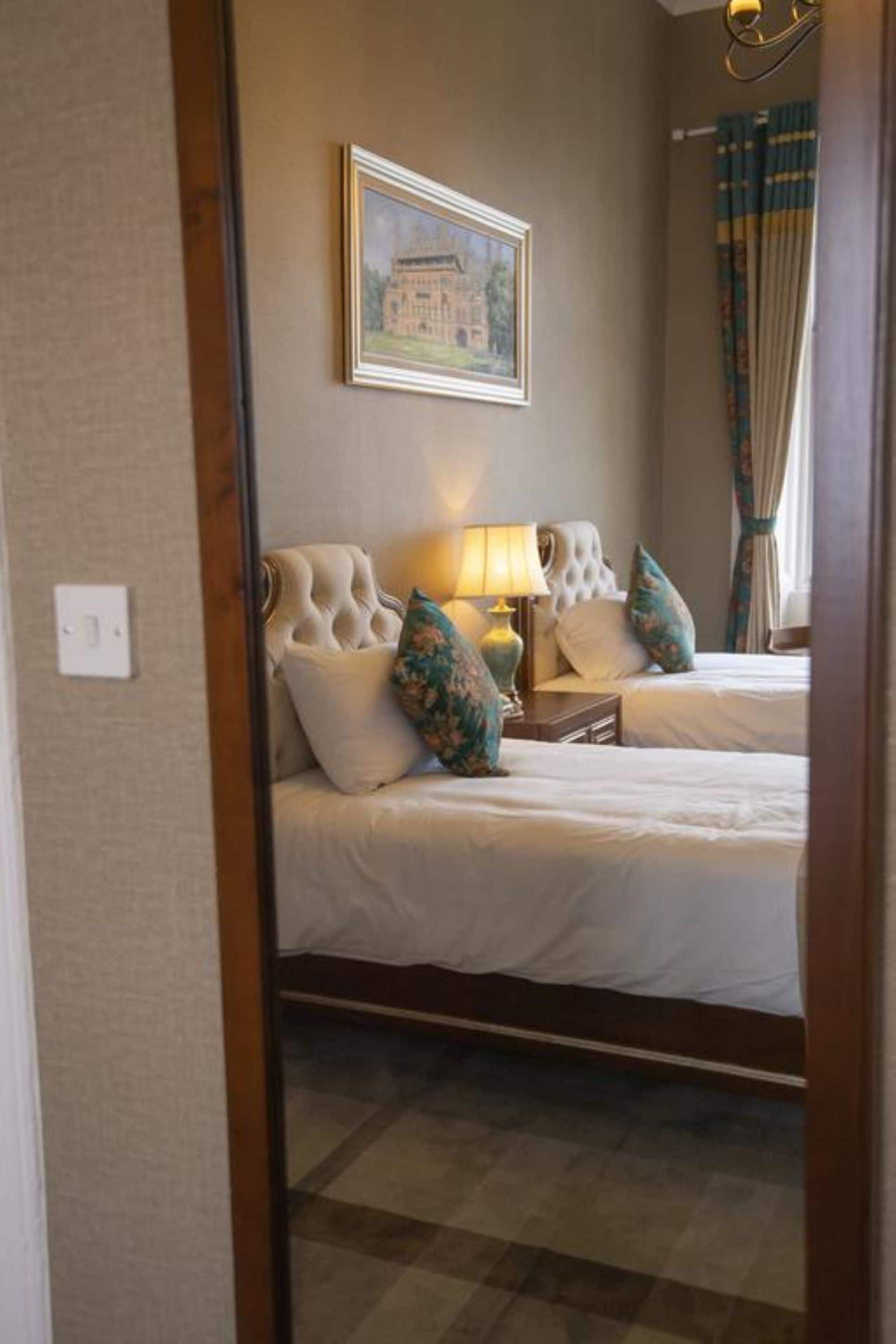 Une chambre d’hôtel avec deux lits, un tableau encadré au mur, une lampe et une fenêtre avec des rideaux.