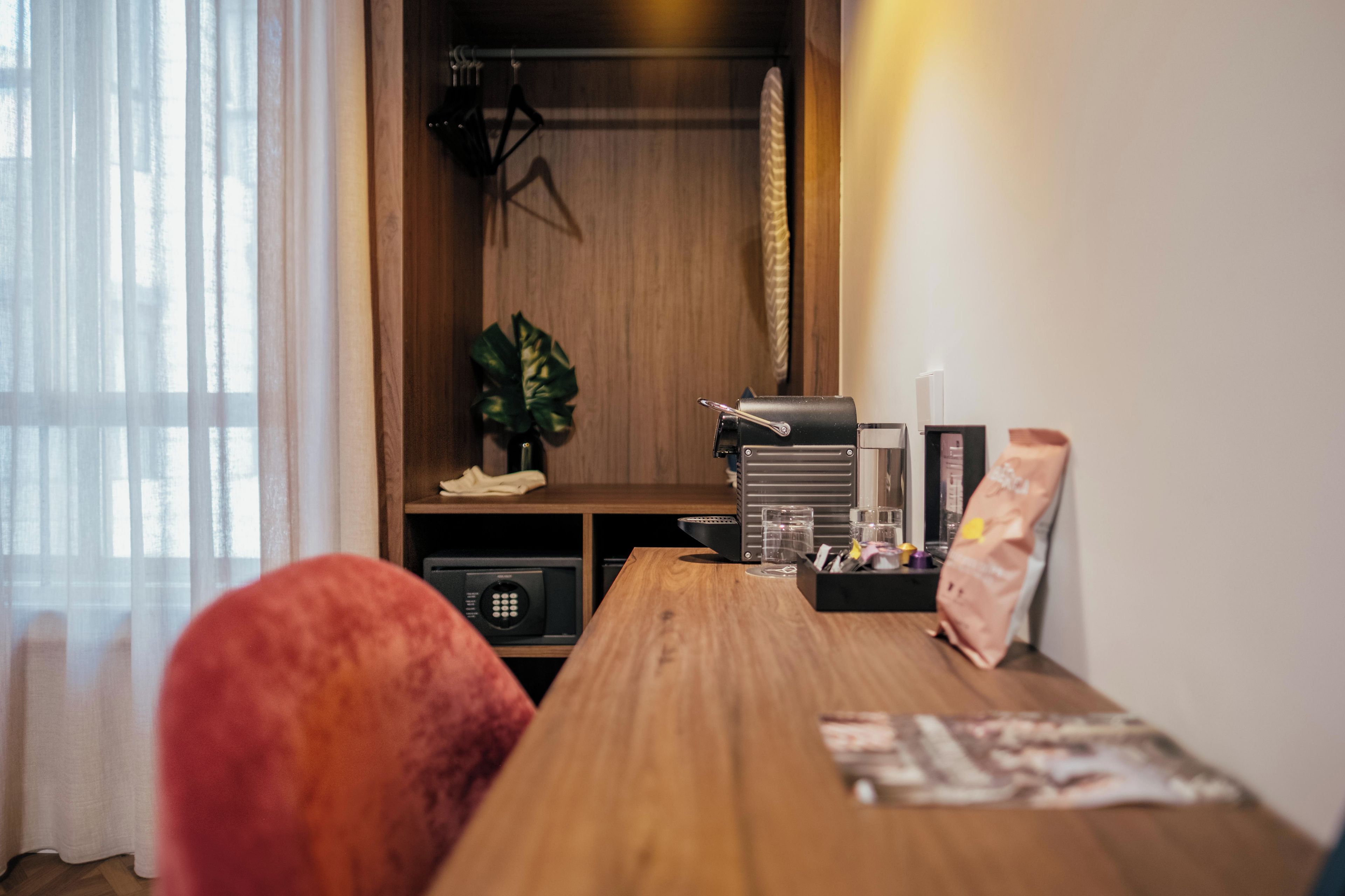 A hotel room with a wooden table, a red chair, a plant, a minibar, and a window with curtains.