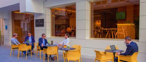 A group of people seated at outdoor tables with yellow chairs, outside a building with large windows and a sign that reads "Hotel".