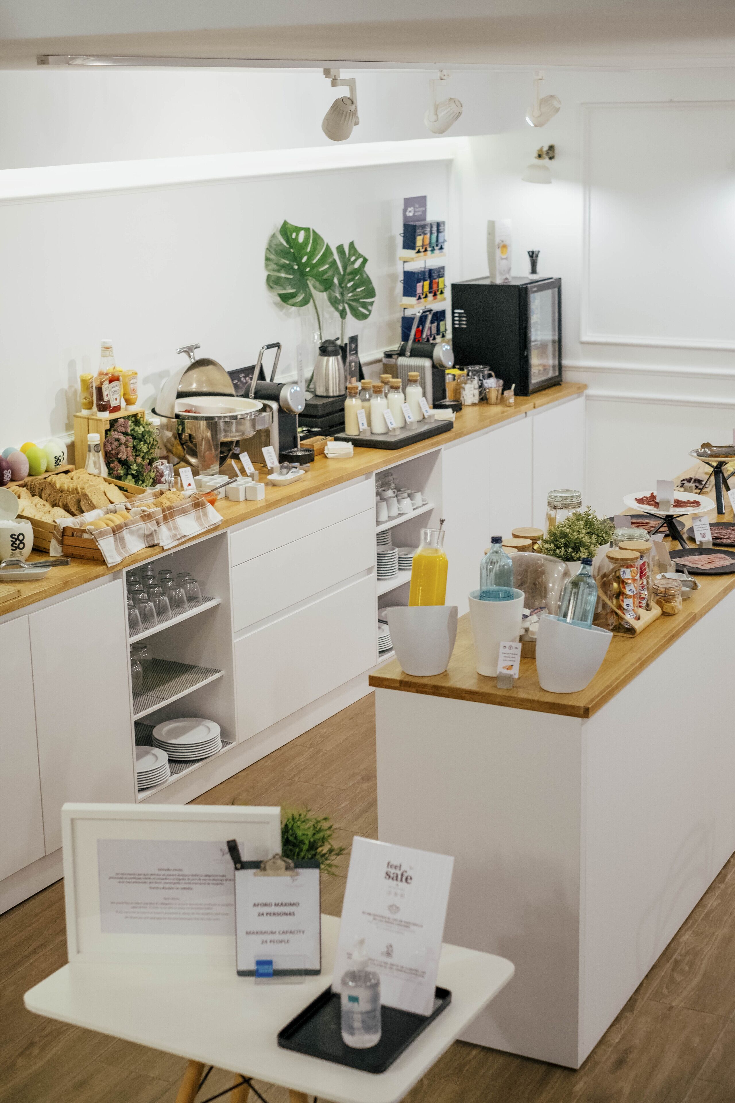 Una barra de desayuno con diversos alimentos y bebidas, un mostrador blanco con electrodomésticos de cocina y un letrero que indica una capacidad máxima de 24 personas.