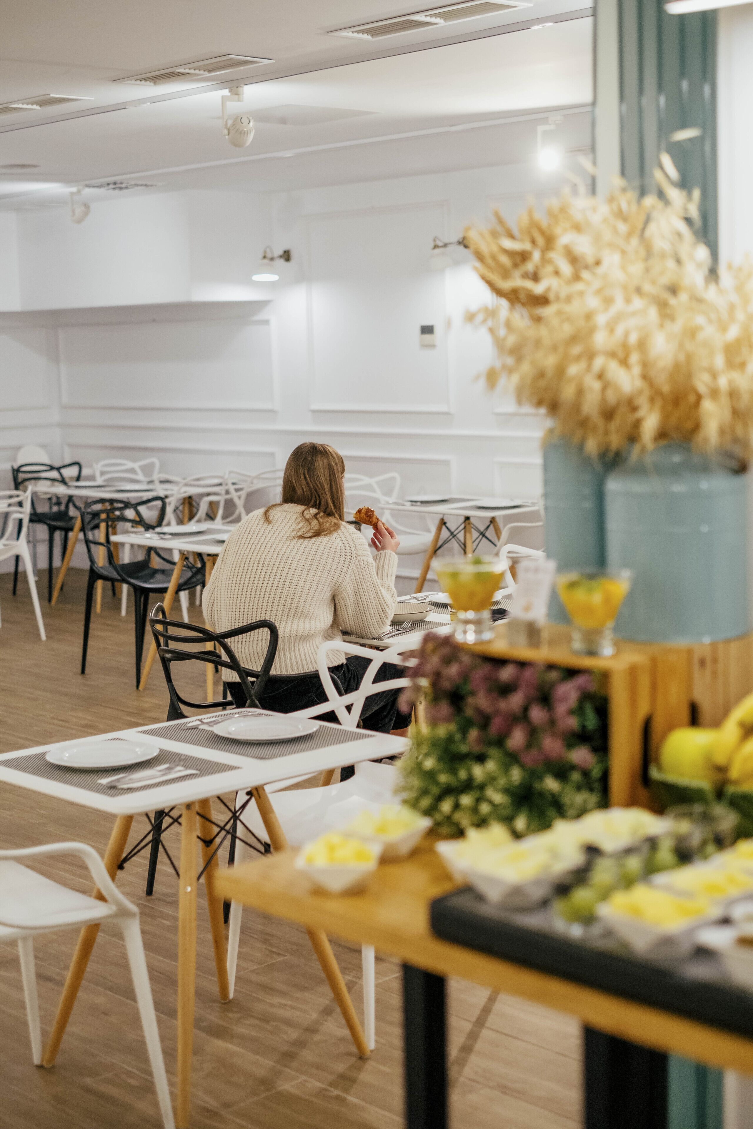 A dining area with white tables and chairs, a person seated at a table with a flower arrangement and a drink.
