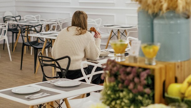 Comedor con mesas y sillas blancas, una persona sentada en una mesa con un arreglo floral y una bebida.