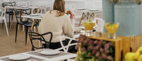 Comedor con mesas y sillas blancas, una persona sentada en una mesa con un arreglo floral y una bebida.